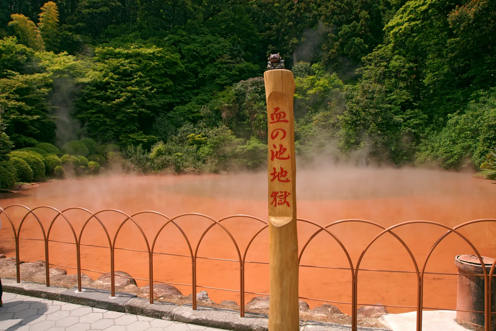 冒著蒸氣的紅色池子,前方是圓弧鐵欄杆和寫著「血の池地獄」的木柱