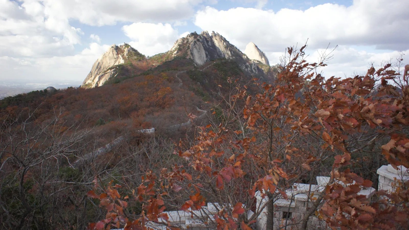 The Bukhansanseong fortress wall running towards the granite peaks, with rust-red oak leaves in the foreground