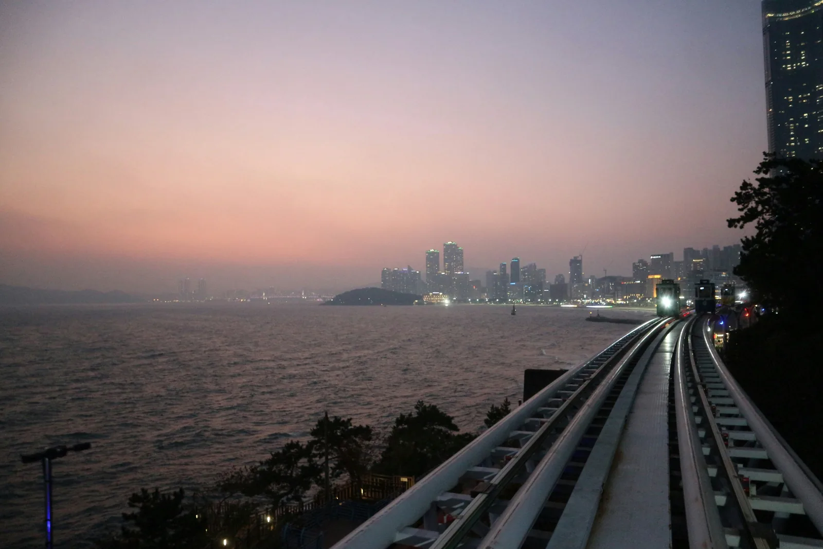 黃昏時並排的天空膠囊列車軌道和亮著燈的膠囊,海的對面是海雲台的高樓和冬柏島