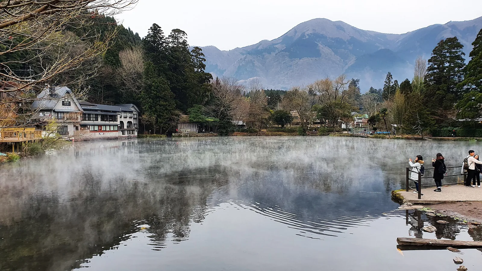寒冷早晨，金鱗湖湖面升起白霧，湖畔有建築與落葉的樹木，遠方是山巒，幾位遊客站在小觀景台上