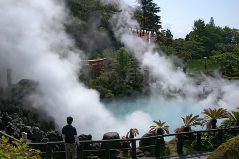 Steam pouring off the sea-blue pool at Umi Jigoku, with a red torii in the garden and a visitor standing at the railing
