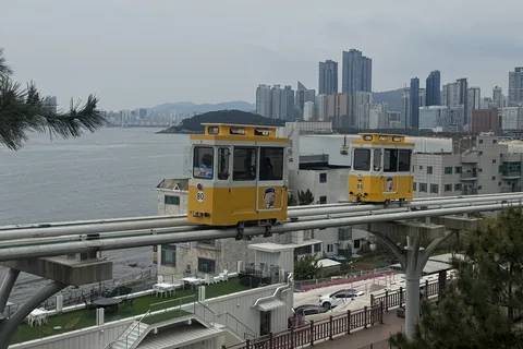 Two yellow Sky Capsules on an elevated coastal track, with Haeundae's high-rises and the bay behind