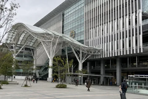 A broad paved plaza beneath a branching white canopy outside a large transit building
