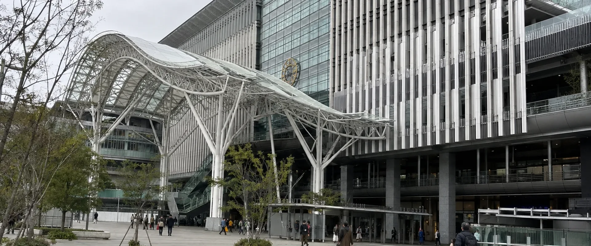 A broad paved plaza beneath a branching white canopy outside a large transit building