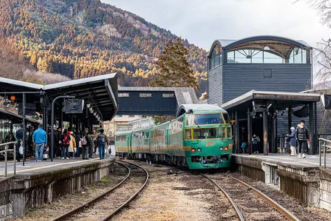 The green Yufuin no Mori train pulling into Yufuin Station, with the black wooden station building and a forested hillside behind the platforms