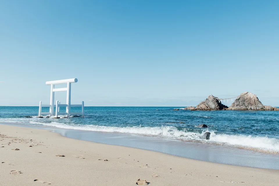 A white torii standing in the shallows off the sandy beach at Futamigaura, with the two Meoto-iwa rocks joined by a rope to the right