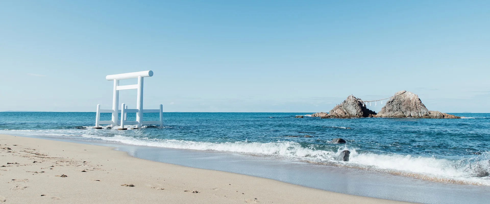 A white torii standing in the shallows off the sandy beach at Futamigaura, with the two Meoto-iwa rocks joined by a rope to the right