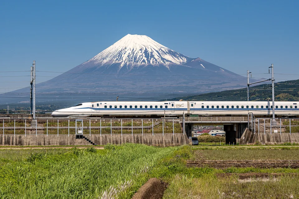 A Tokaido Shinkansen N700S train passing below snow-capped Mount Fuji, with fields in the foreground