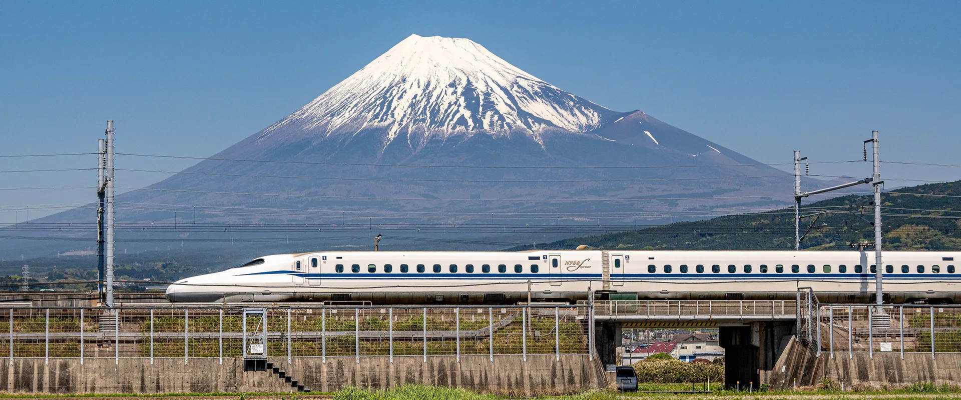 A Tokaido Shinkansen N700S train passing below snow-capped Mount Fuji, with fields in the foreground