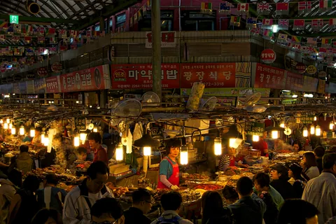 Gwangjang Market's food crossroads at night, with lit stall counters, steam and people passing