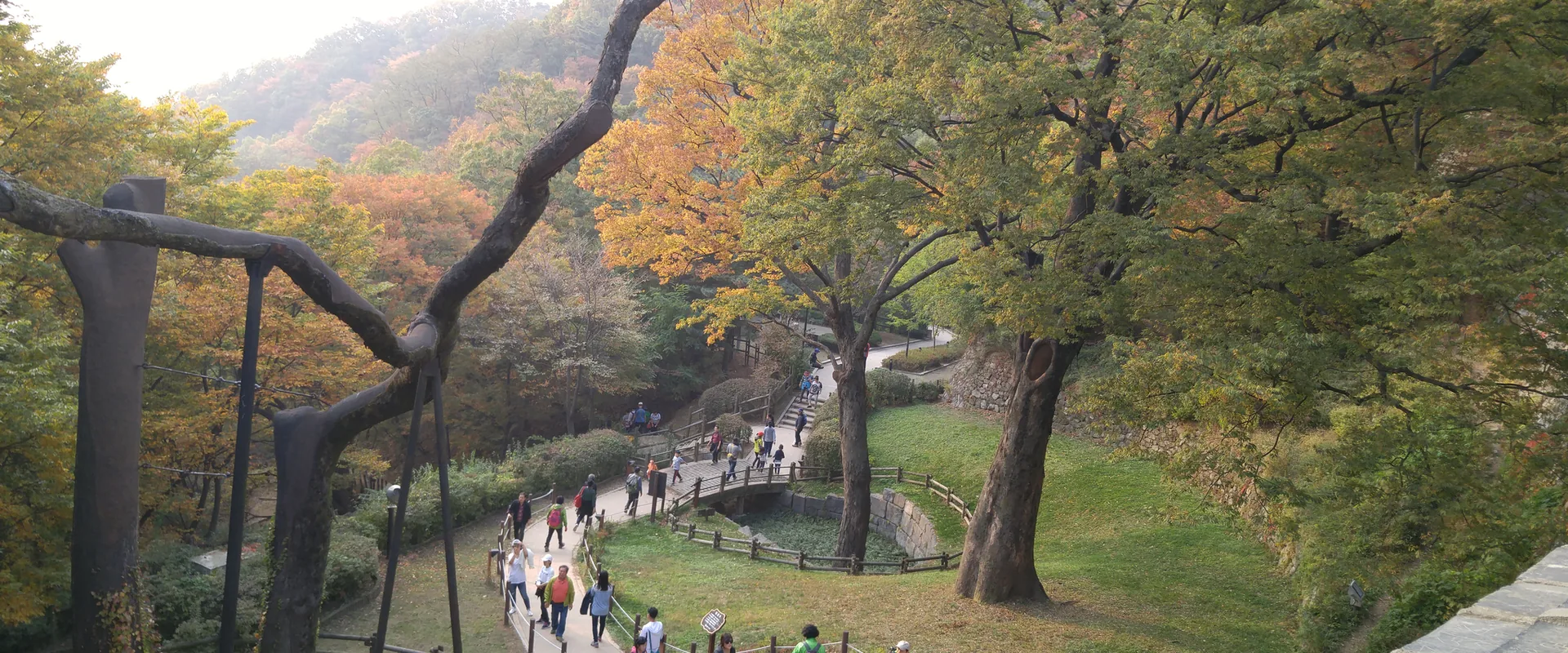 Walkers on a winding path below the South Gate of Namhansanseong, among trees turning orange and yellow