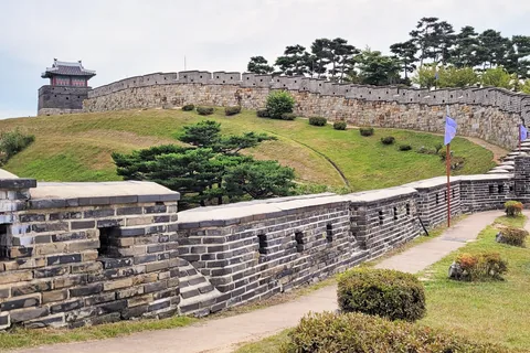 The stone wall of Hwaseong Fortress curving over a grassy rise to a watchtower, with the wall-top path in front