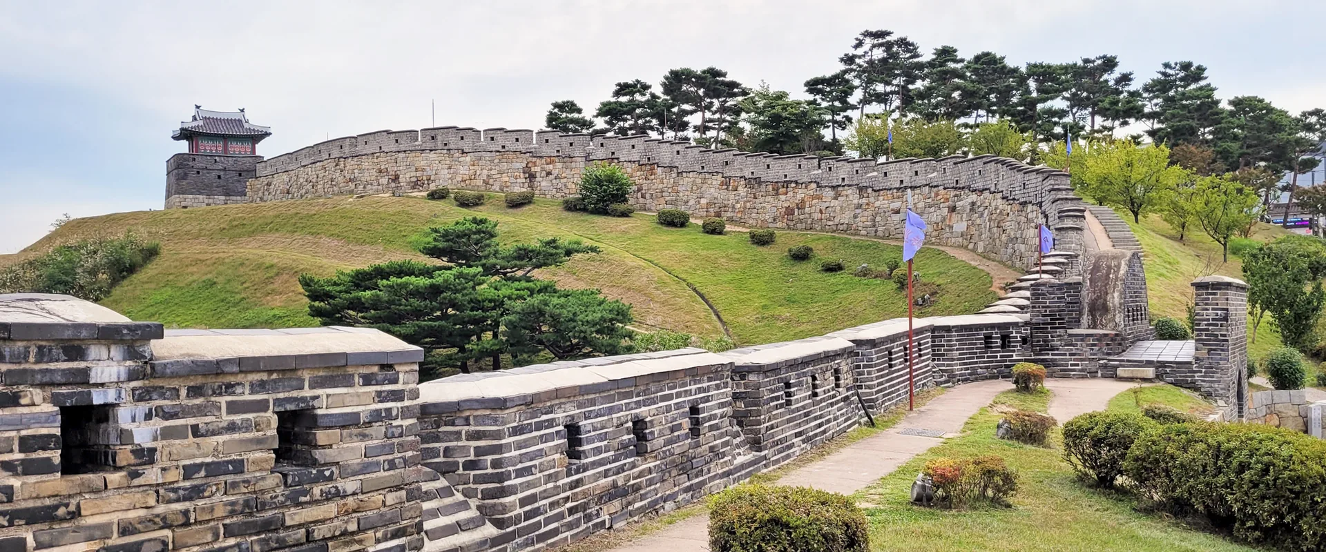 The stone wall of Hwaseong Fortress curving over a grassy rise to a watchtower, with the wall-top path in front