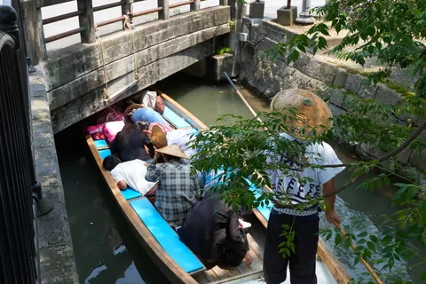 Passengers bend low in a Yanagawa canal boat to pass under a low stone bridge while the boatman in a straw hat stands at the stern