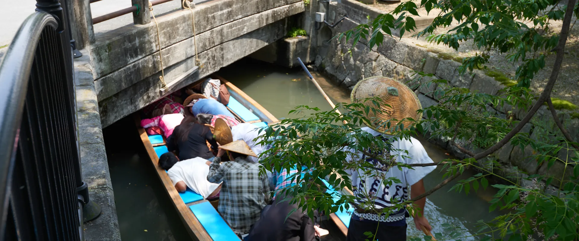 Passengers bend low in a Yanagawa canal boat to pass under a low stone bridge while the boatman in a straw hat stands at the stern