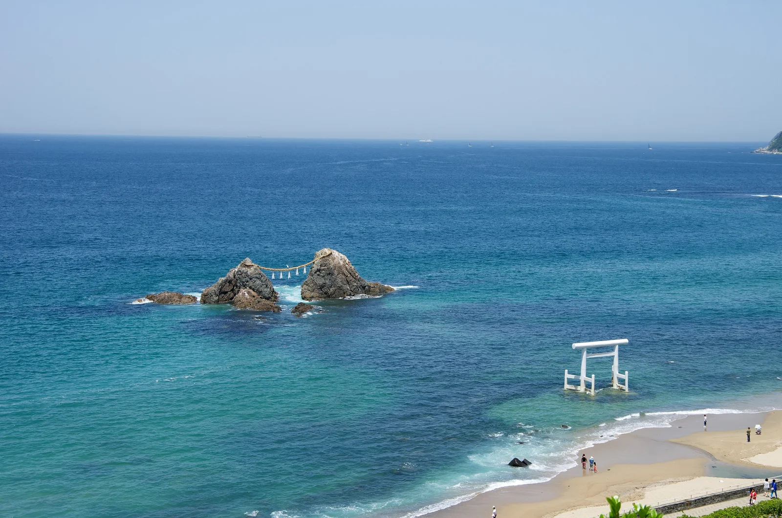 Futamigaura seen from the hillside: two rocks joined by a rope in blue water, and a white torii standing in the shallows near the sand