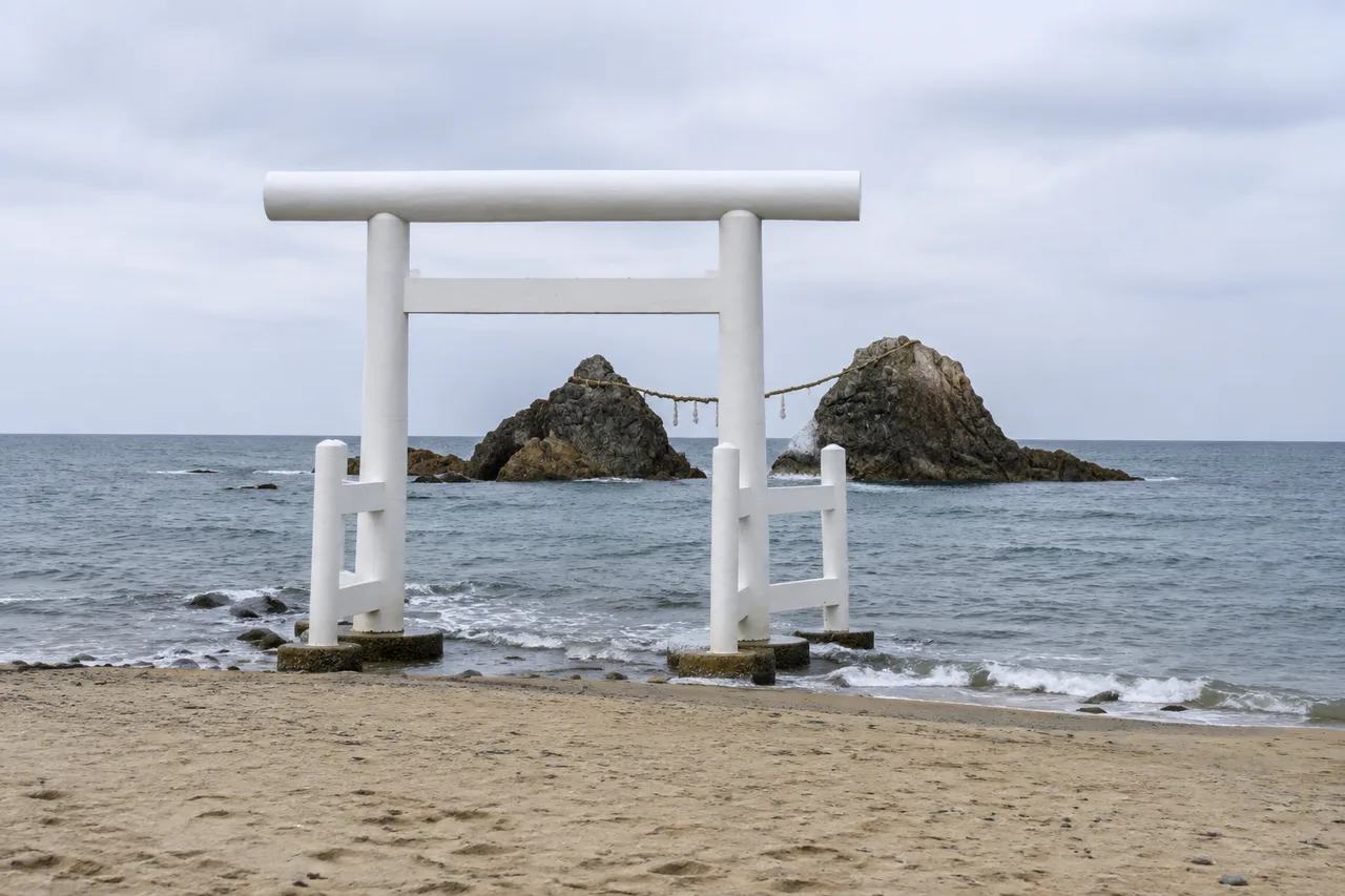 A complete white torii on the beach, with two offshore rocks joined by a shimenawa rope behind it.
