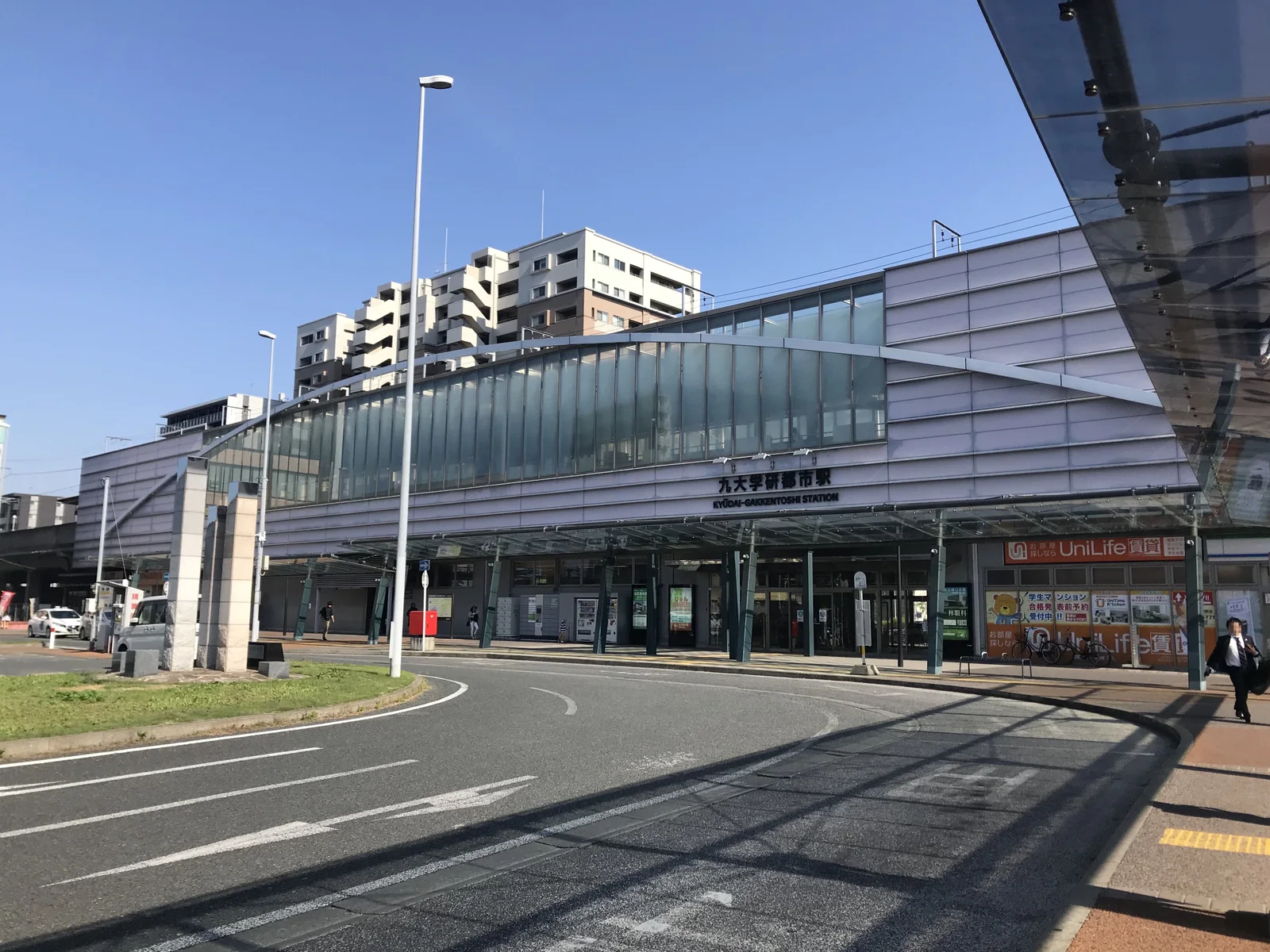 Kyudai-Gakkentoshi Station building with a long glass canopy and the bus and taxi bays in front