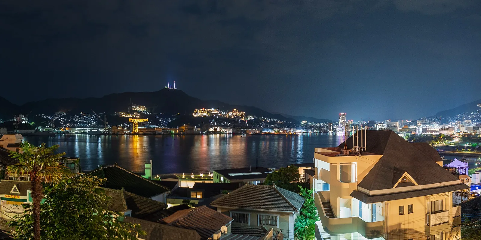 Nagasaki Harbour and the hills across the water at night, seen over the roofs of Western-style houses on a hillside