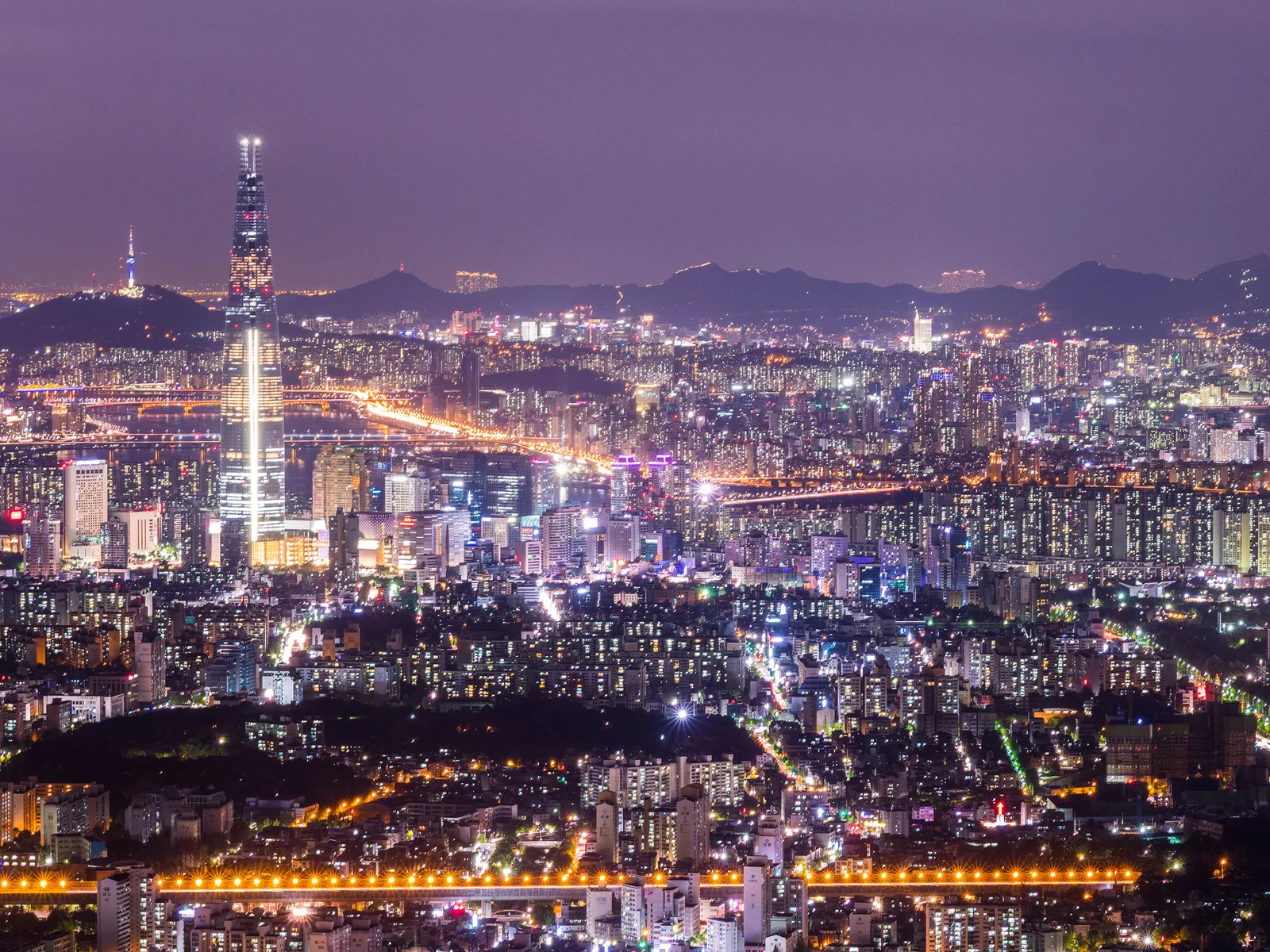 Seoul lit up at night, with Lotte World Tower on the left and mountains along the horizon