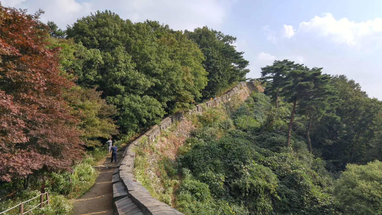 A dirt path running beside the stone fortress wall, with a red-leaved maple on the left and walkers ahead