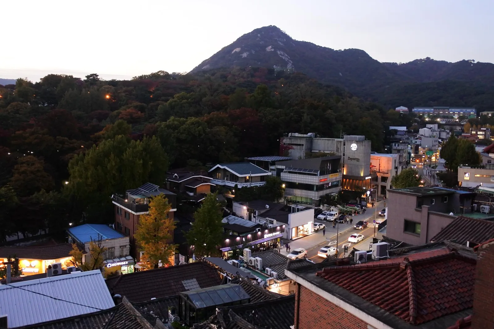 Samcheong-dong at dusk seen from above, with lit shop fronts, cars on the street, trees turning colour and Bugaksan behind