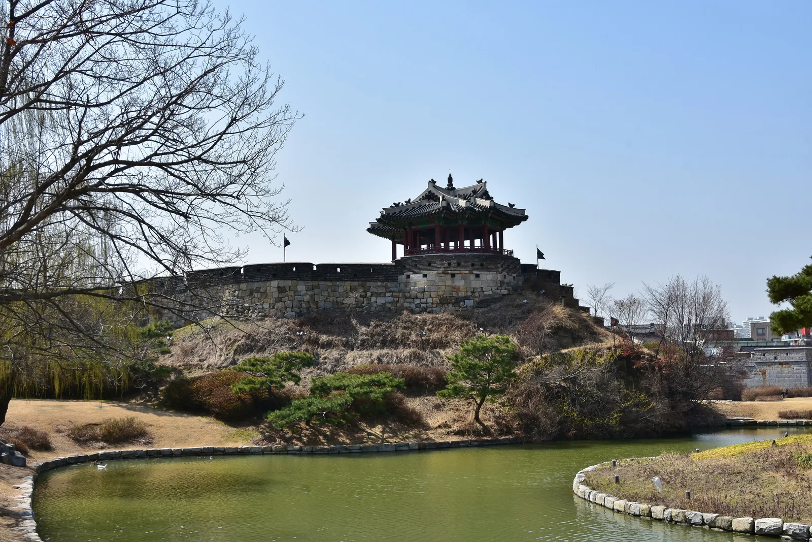 Banghwasuryujeong pavilion on a mound above a pond, joined to the fortress wall, with bare trees around it