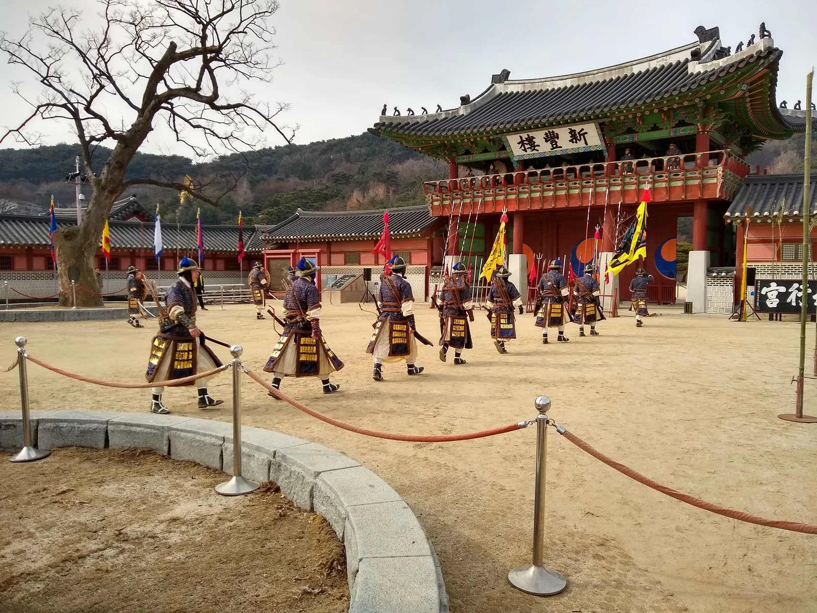 A line of people in Joseon-era military costume walking across the sandy forecourt in front of Sinpungnu, the main gate of Hwaseong Haenggung
