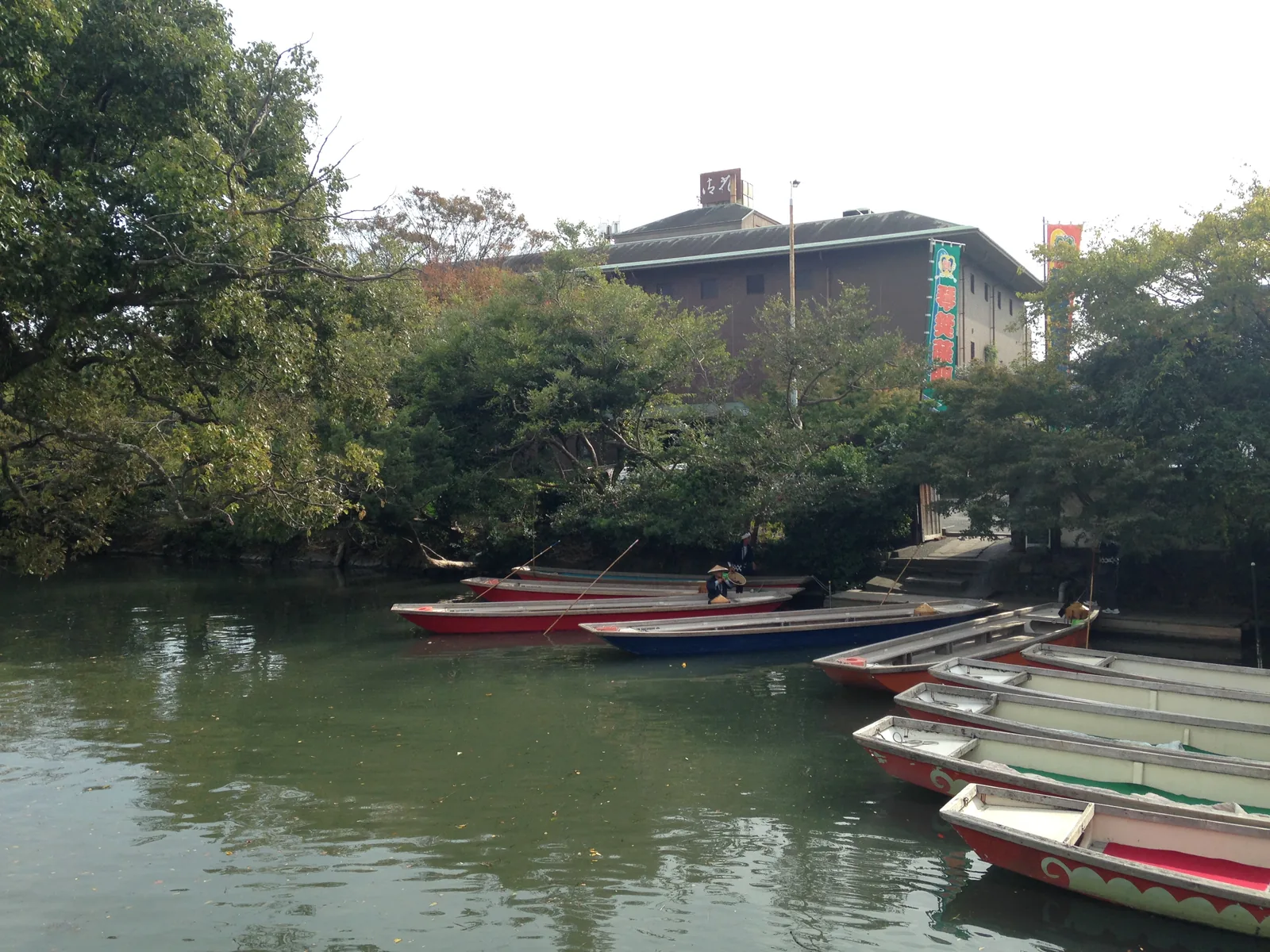 A row of canal boats tied up along a tree-lined bank, with buildings behind