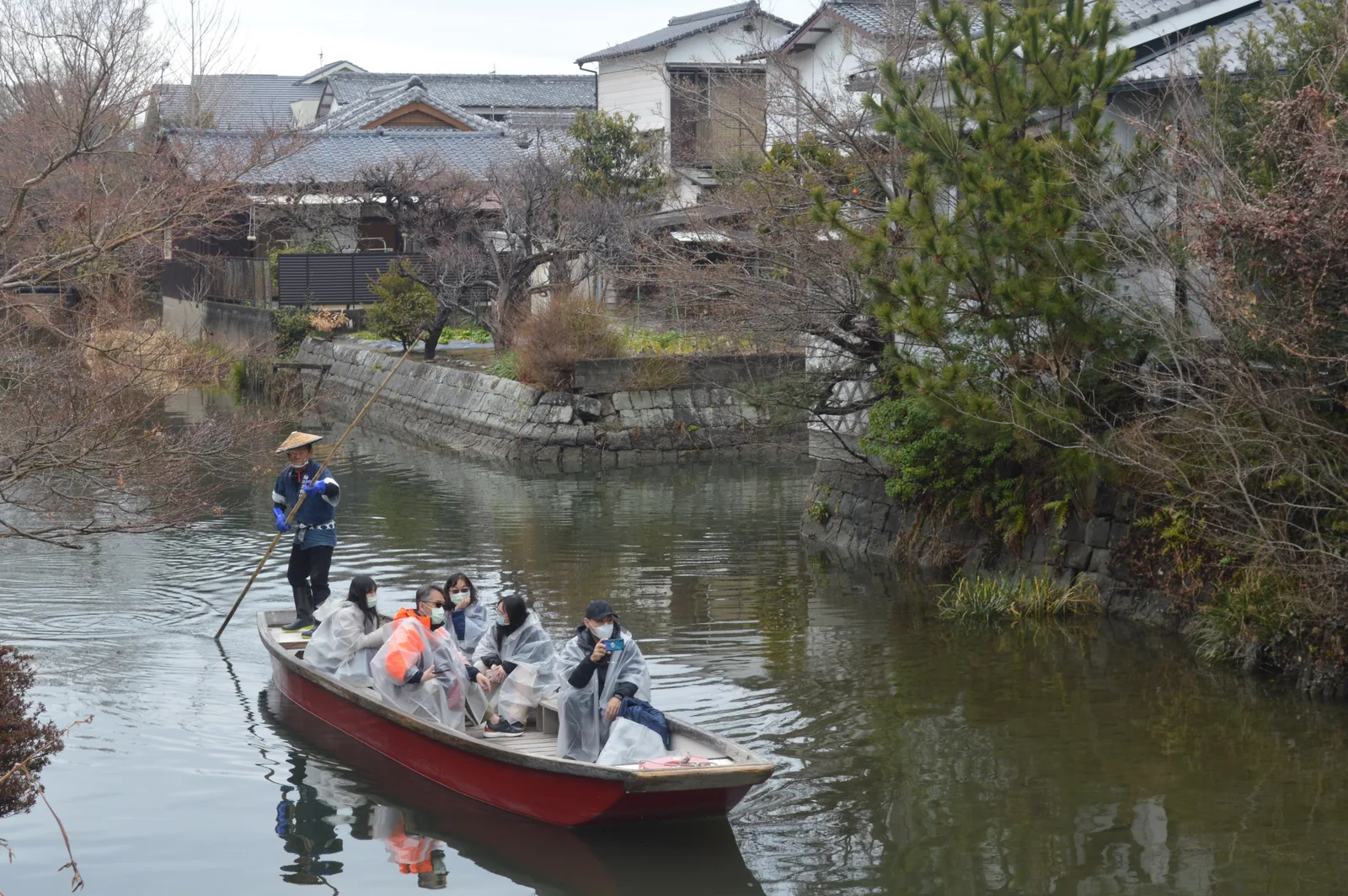 Passengers in clear plastic ponchos ride a canal boat down a narrow waterway while the boatman poles from the back